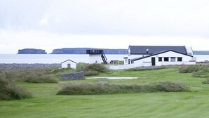 A coastal house with a grassy lawn, a small adjacent building, and cliffs in the background. - Lónkot Rural Resort (Skagafjörour)