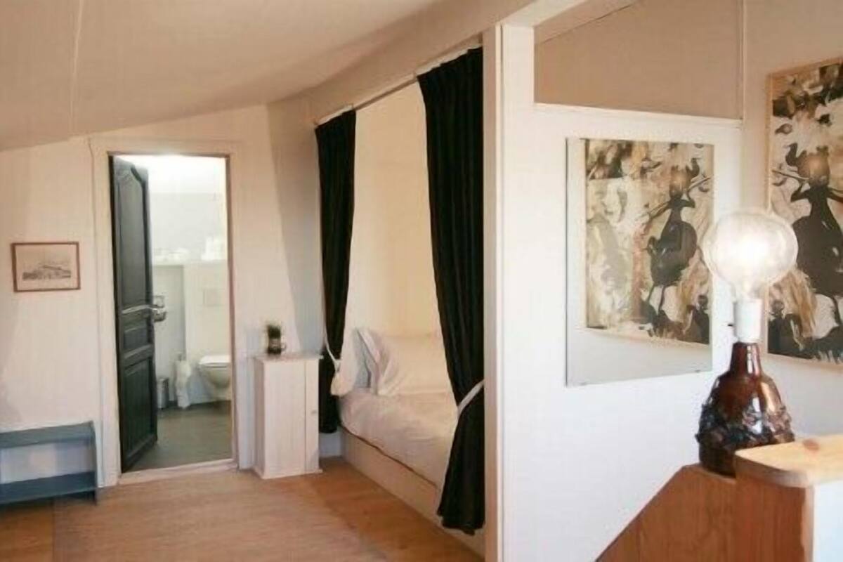 A hotel room with a bed, a small bedside table, a nightstand, a painting on the wall, and a door leading to the bathroom.