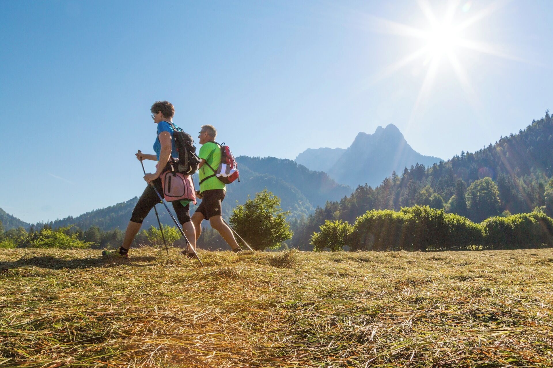 Two hikers walking on a grassy field with a mountainous backdrop and a clear sky.