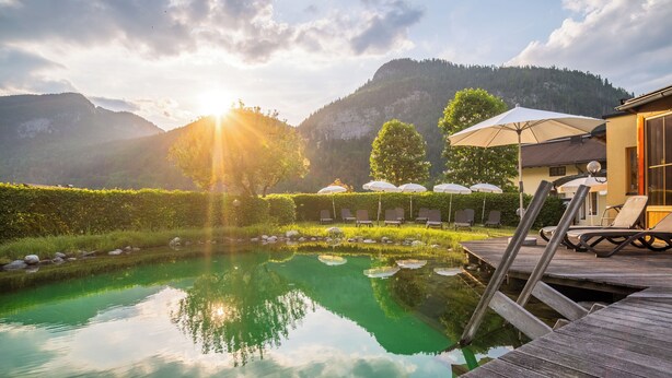 A serene outdoor pool area with a wooden deck, lounge chairs, and umbrellas, set against a backdrop of mountains and a clear sky. - Hotel Vitaloase zu den drei Brüdern (Unken)