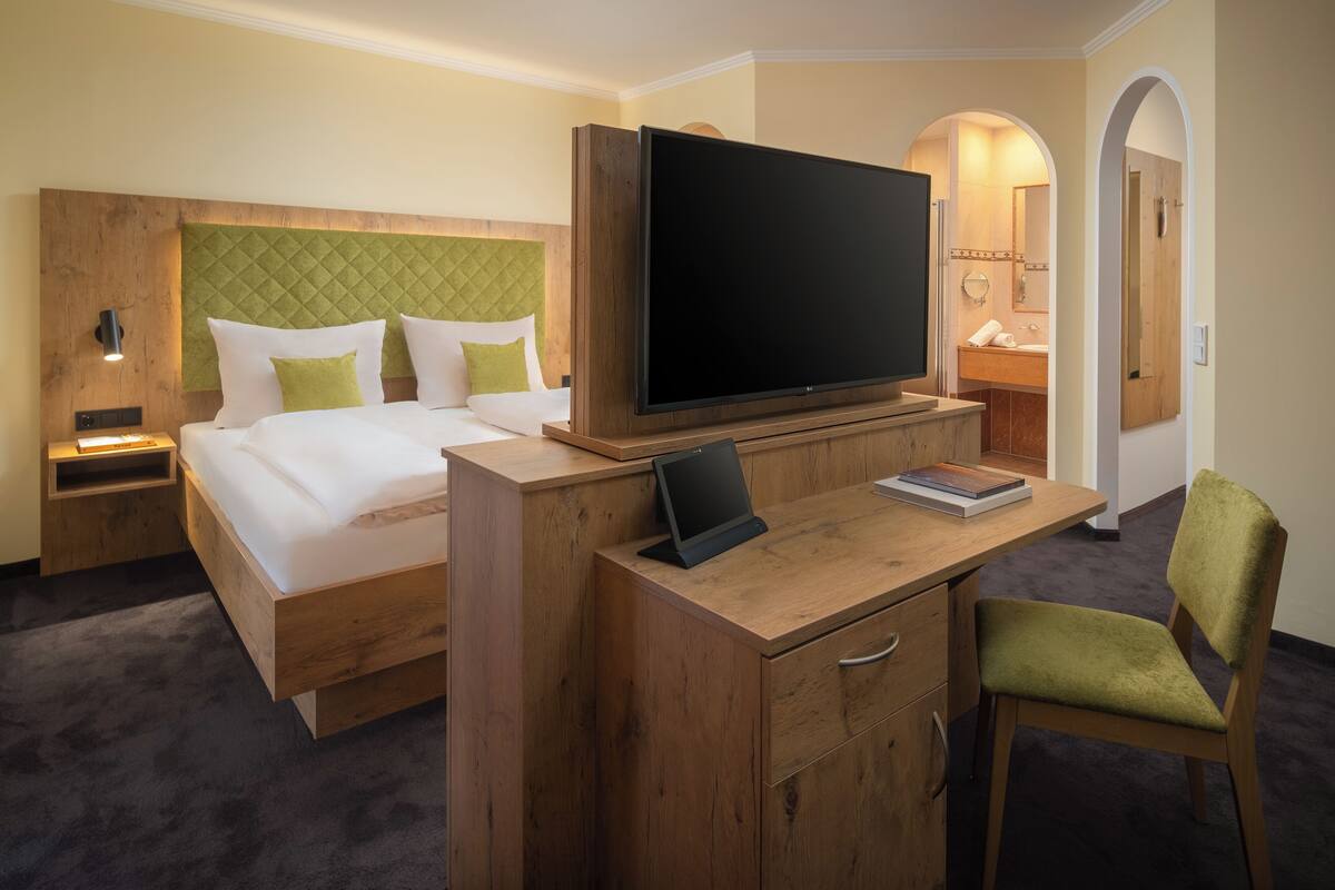 Ein Hotelzimmer mit einem Bett, einem Schreibtisch mit Flachbildfernseher, einem Sessel und einem durch einen Bogen erreichbaren Badezimmer.