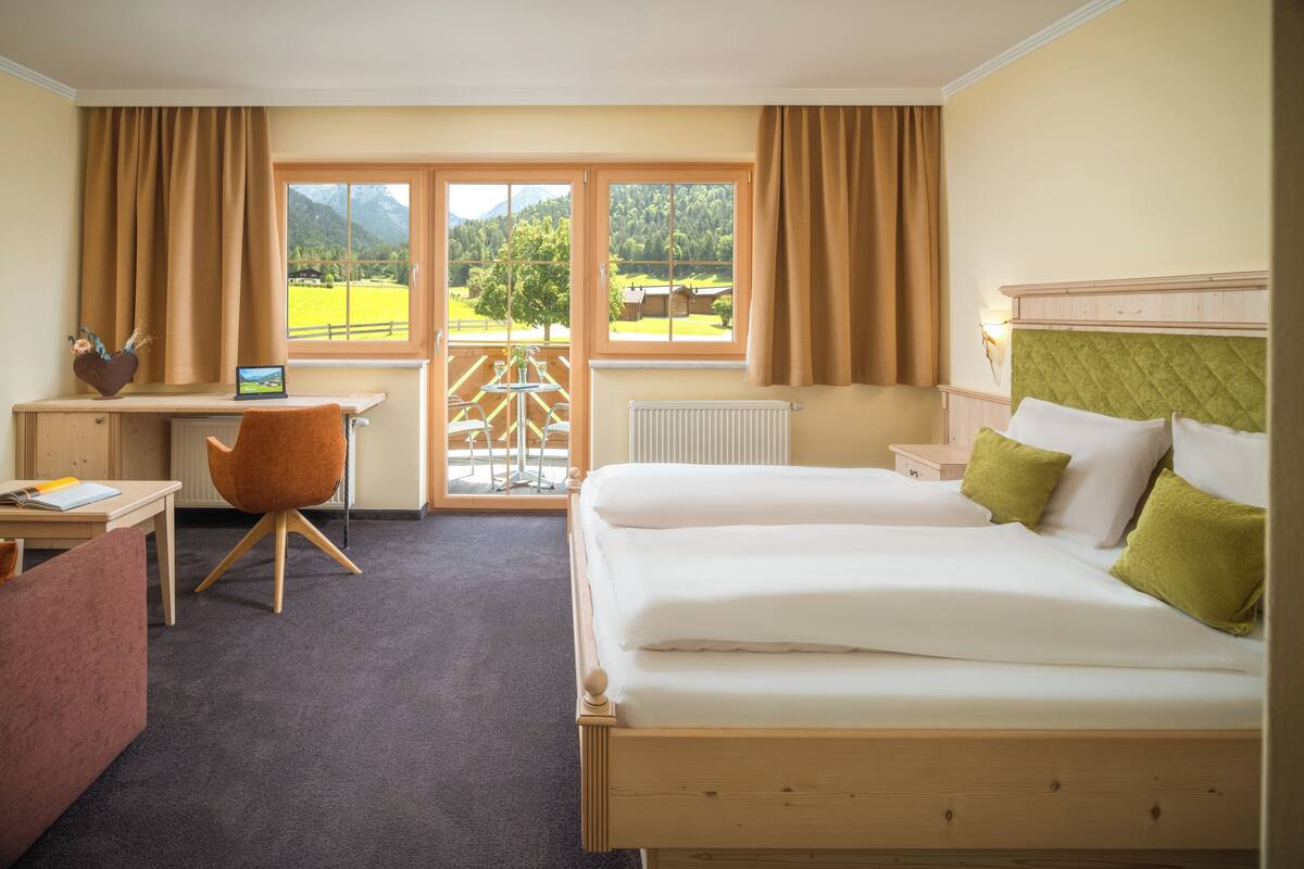 Ein Hotelzimmer mit einem hölzernen Bett, einem Schreibtisch mit Stuhl, Blick auf eine grüne Landschaft durch große Fenster und einem Heizkörper an der Wand.