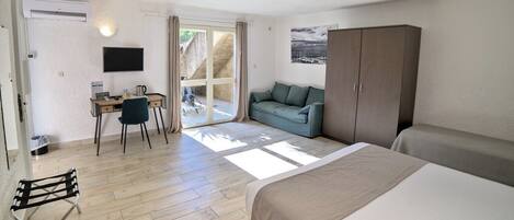 A room with a bed, a desk with a chair, a sofa, and a wardrobe. There is a sliding glass door leading to an outdoor area.