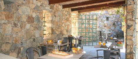Un espace extérieur aménagé pour les repas, avec des murs en pierre, des chaises et des tables en métal, le tout préparé pour le petit-déjeuner.