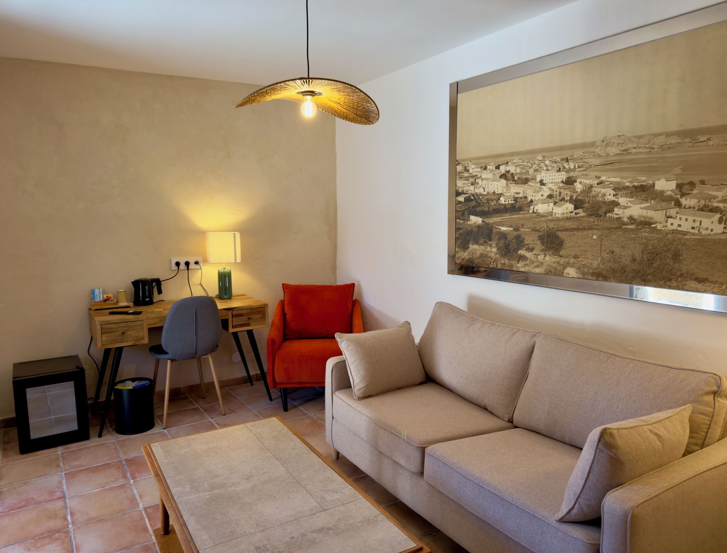 A living room with a sofa, a desk with a lamp, a chair, a coffee maker, and a framed picture on the wall.