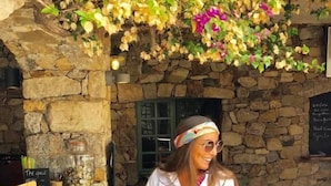 A person seated at a bar with a stone wall backdrop, a chalkboard menu, and a hanging floral arrangement.