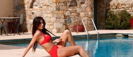 A woman in a red swimsuit sitting by a pool with stone walls and a tiled roof in the background.