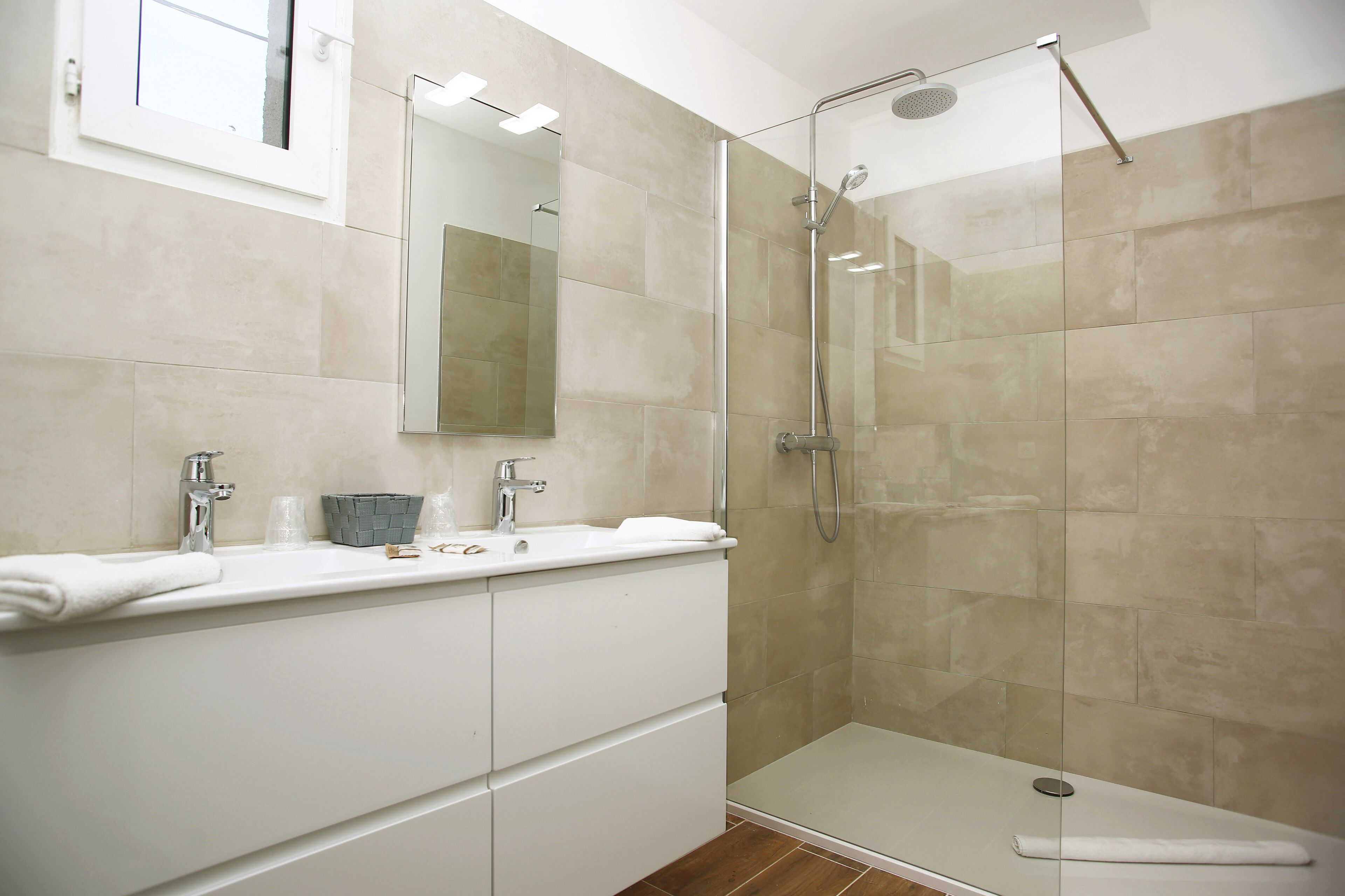 Une salle de bain moderne avec une douche à parois de verre, un meuble-lavabo blanc avec un miroir et des carreaux de couleur beige.