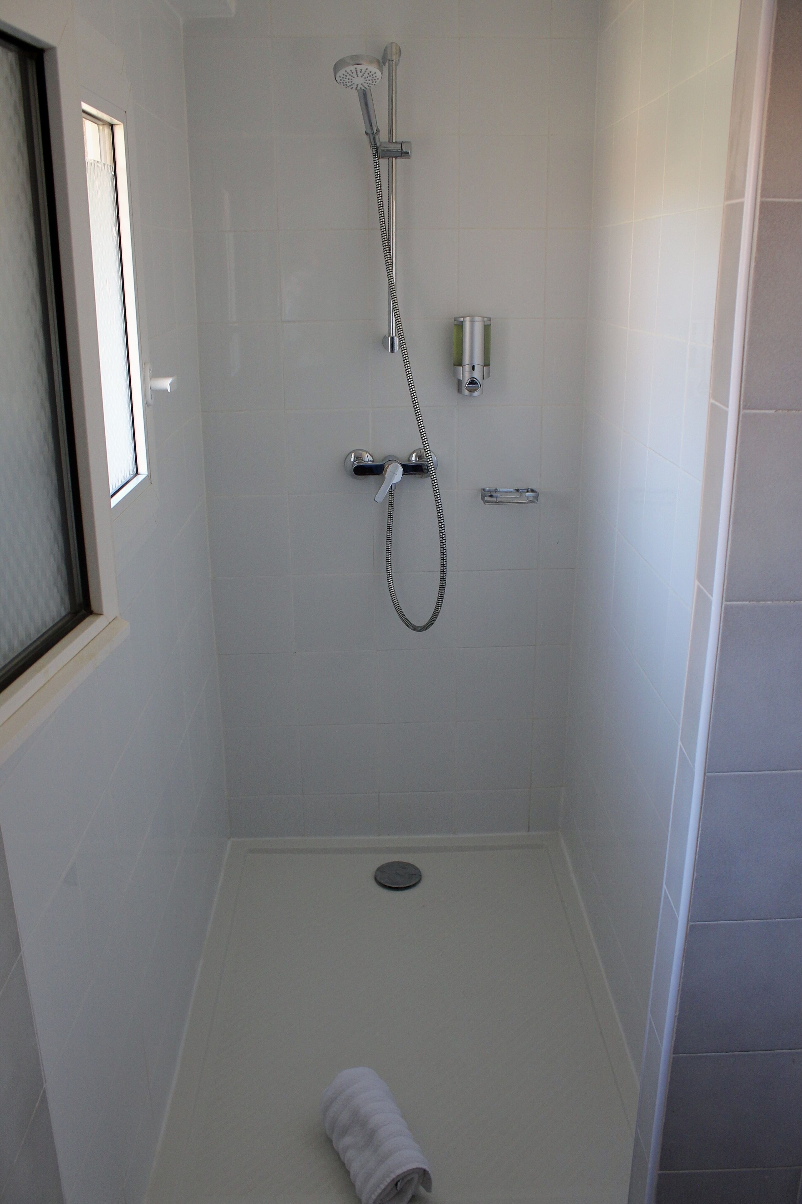 Une petite salle de douche aux murs recouverts de carreaux blancs, équipée d’un pommeau de douche à main, d’un robinet mural et d’une petite étagère.