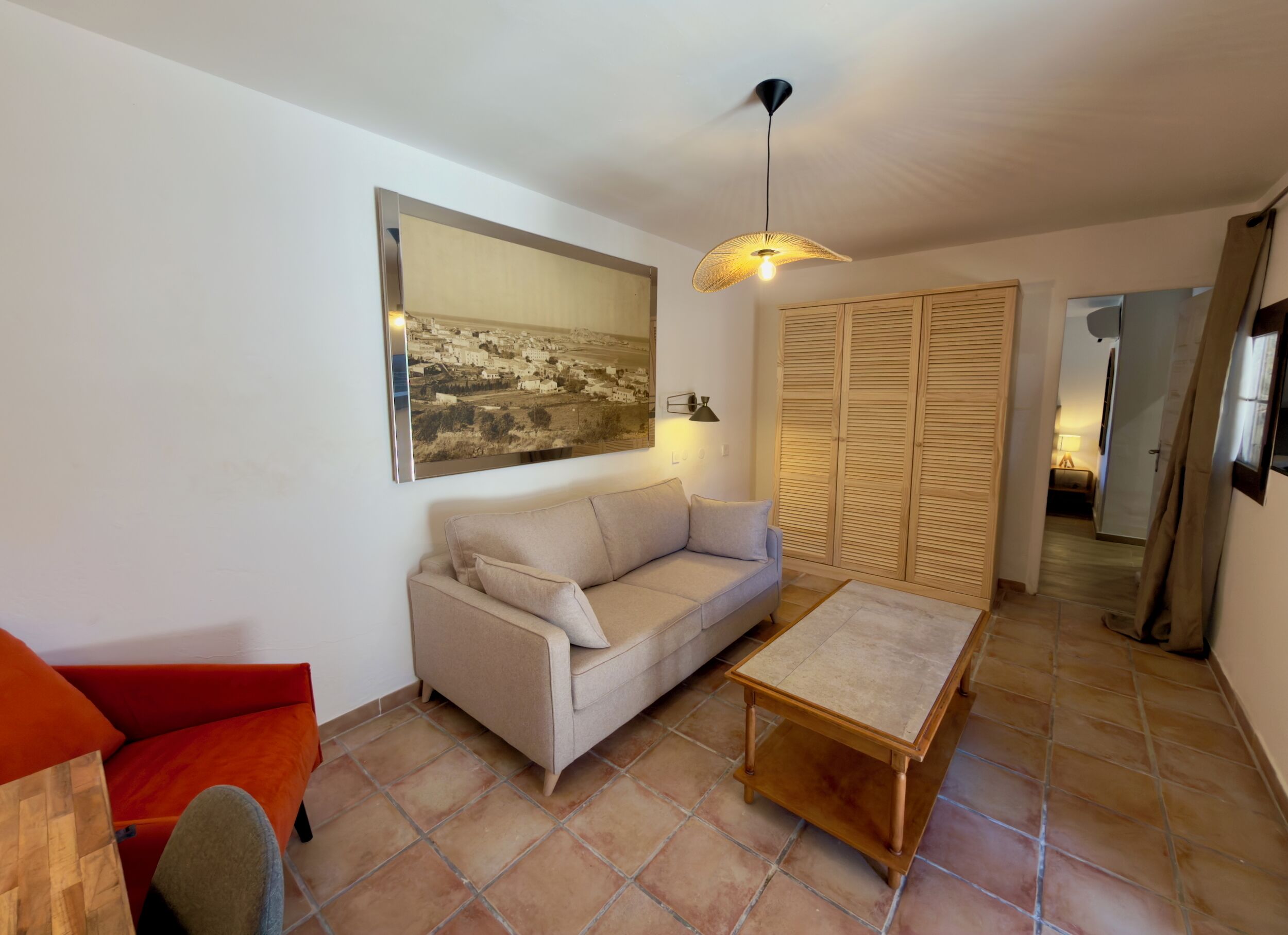 A living room with a sofa, a table, a hanging light fixture, a painting, and a wooden wardrobe.