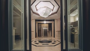 Eine Hotelhalle mit geometrischem Bodenmuster, einem zentralen Kronleuchter und Glastüren zu einem Flur.