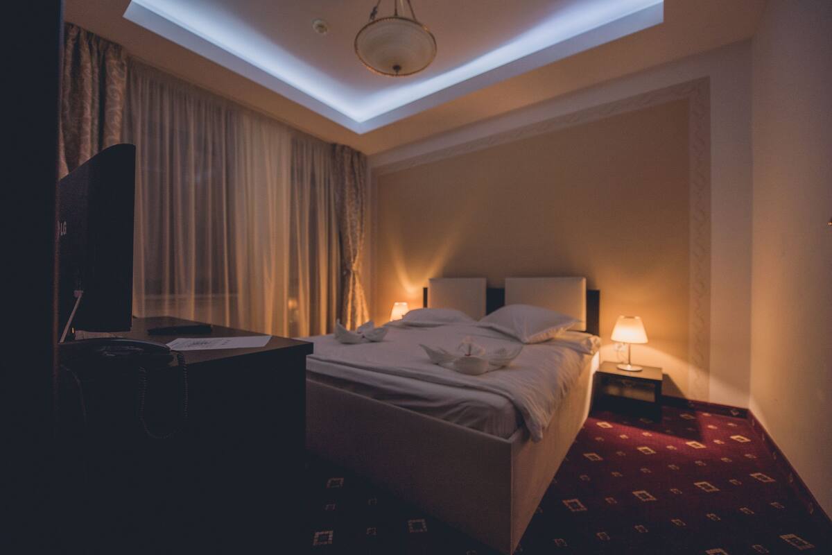 A hotel room with a skylight, a bed with pillows and a decorative pillow, a bedside table with a lamp, and a desk with a chair and a handbag.