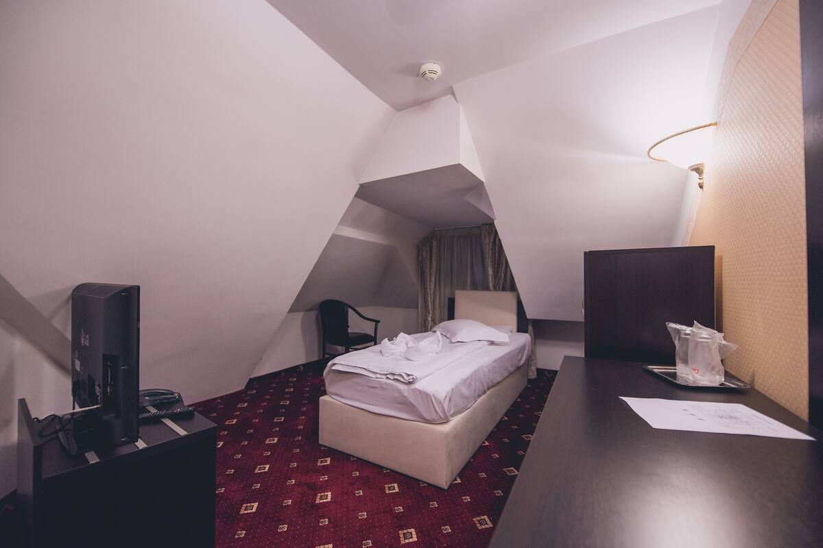 A small, triangular hotel room with a bed, a desk, a chair, and a TV.