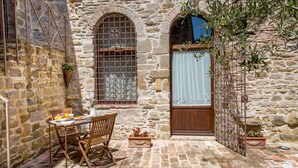 Un bâtiment en pierre avec une table et des chaises en bois, une fenêtre ornée d’une grille métallique et un pot de fleurs.