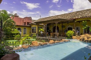 Zona de piscina con terraza cubierta, mobiliario exterior y vista a un edificio amarillo con tejado de azulejos.