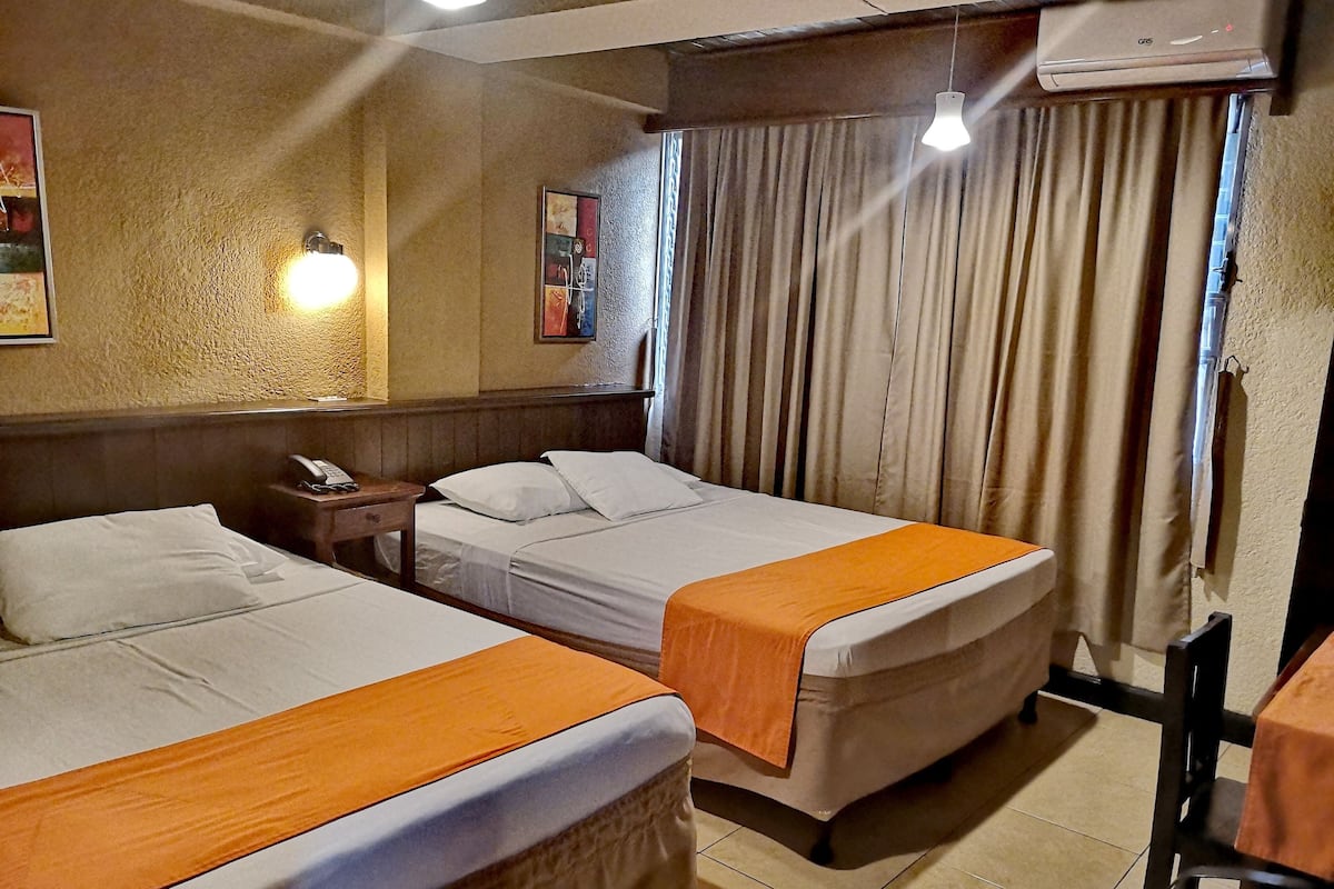 Quarto de hotel com duas camas, uma mesinha de cabeceira com telefone, um quadro na parede e uma janela com cortinas.