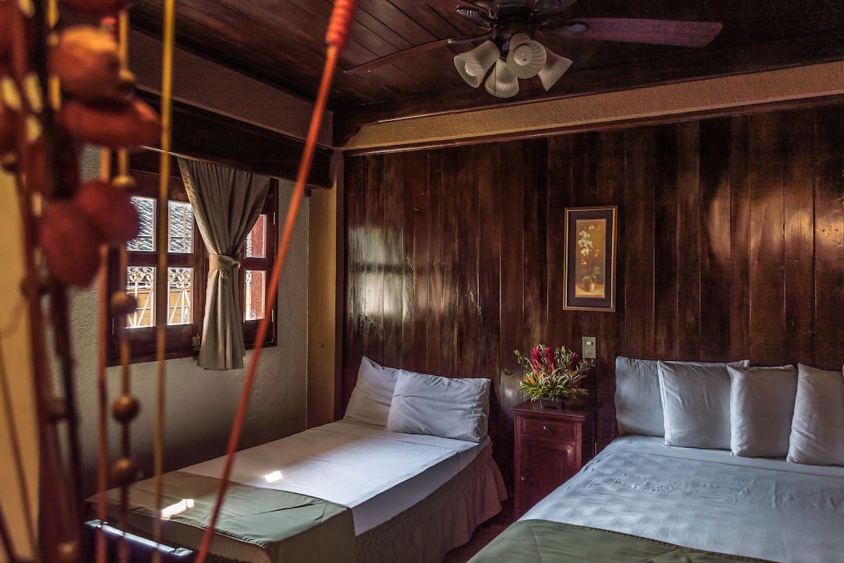 Quarto com duas camas, paredes de madeira, ventoinha no teto, janela com cortinas e uma pequena mesa com um vaso de flores.