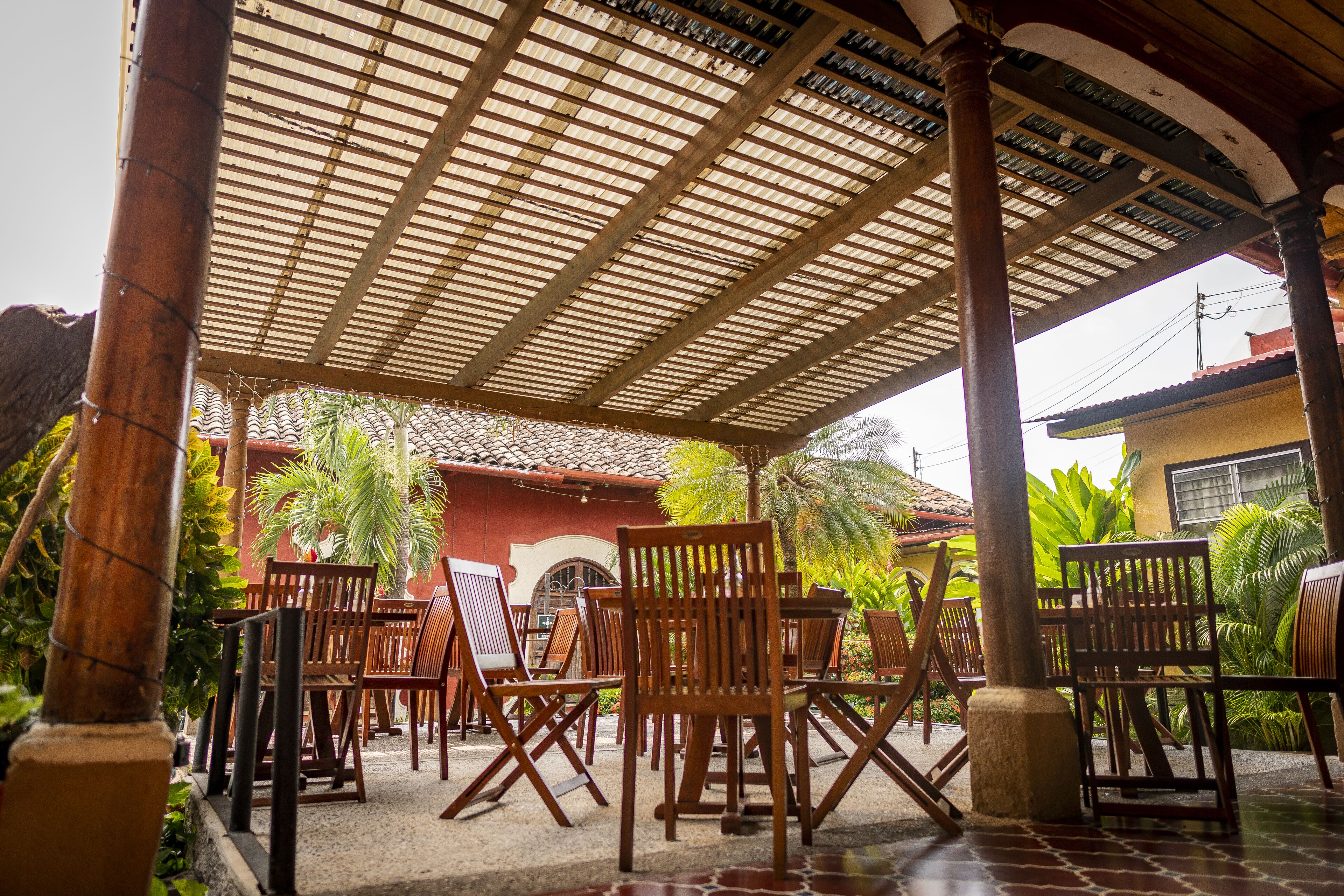 Un espace extérieur aménagé avec des meubles de jardin en bois, comprenant des chaises et des tables, entouré de plantes tropicales et avec, en arrière-plan, un bâtiment doté d’un toit en tuiles.