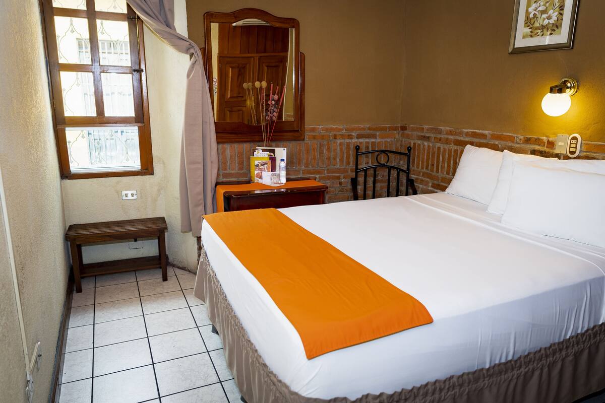 Quarto de hotel com uma cama grande, uma mesinha de cabeceira de madeira, uma pequena mesa, um banco, uma janela com cortinas, um espelho, uma luminária na parede e uma parede de tijolos.