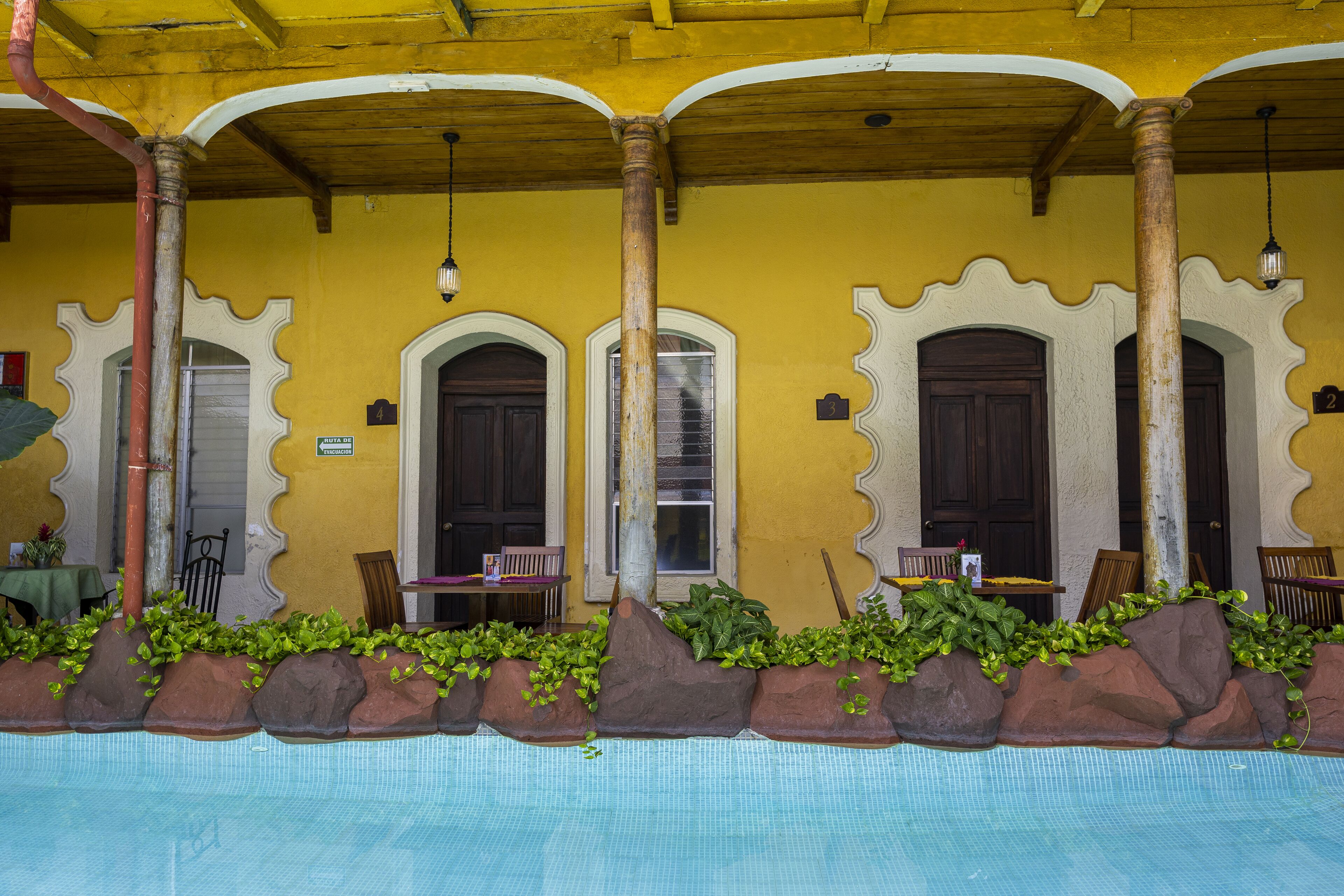 A poolside area with a yellow wall, wooden columns, and a seating area with chairs and tables.