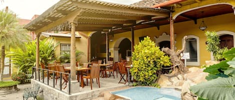 A pool area with a covered patio, outdoor seating, and tropical plants.
