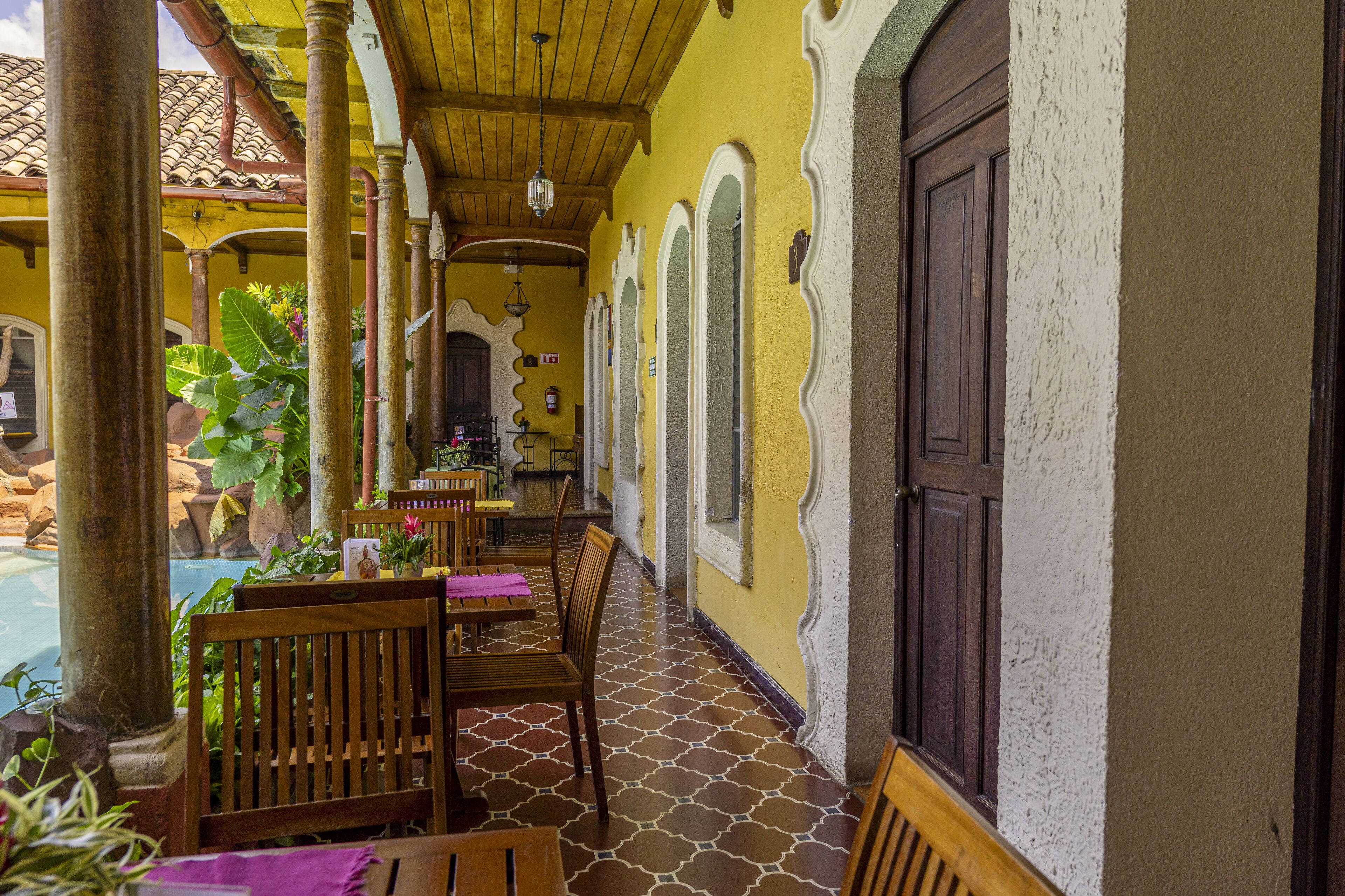 Un espacio comedor al aire libre techado, con mesas y sillas de madera, piso de baldosas y una piscina al fondo.