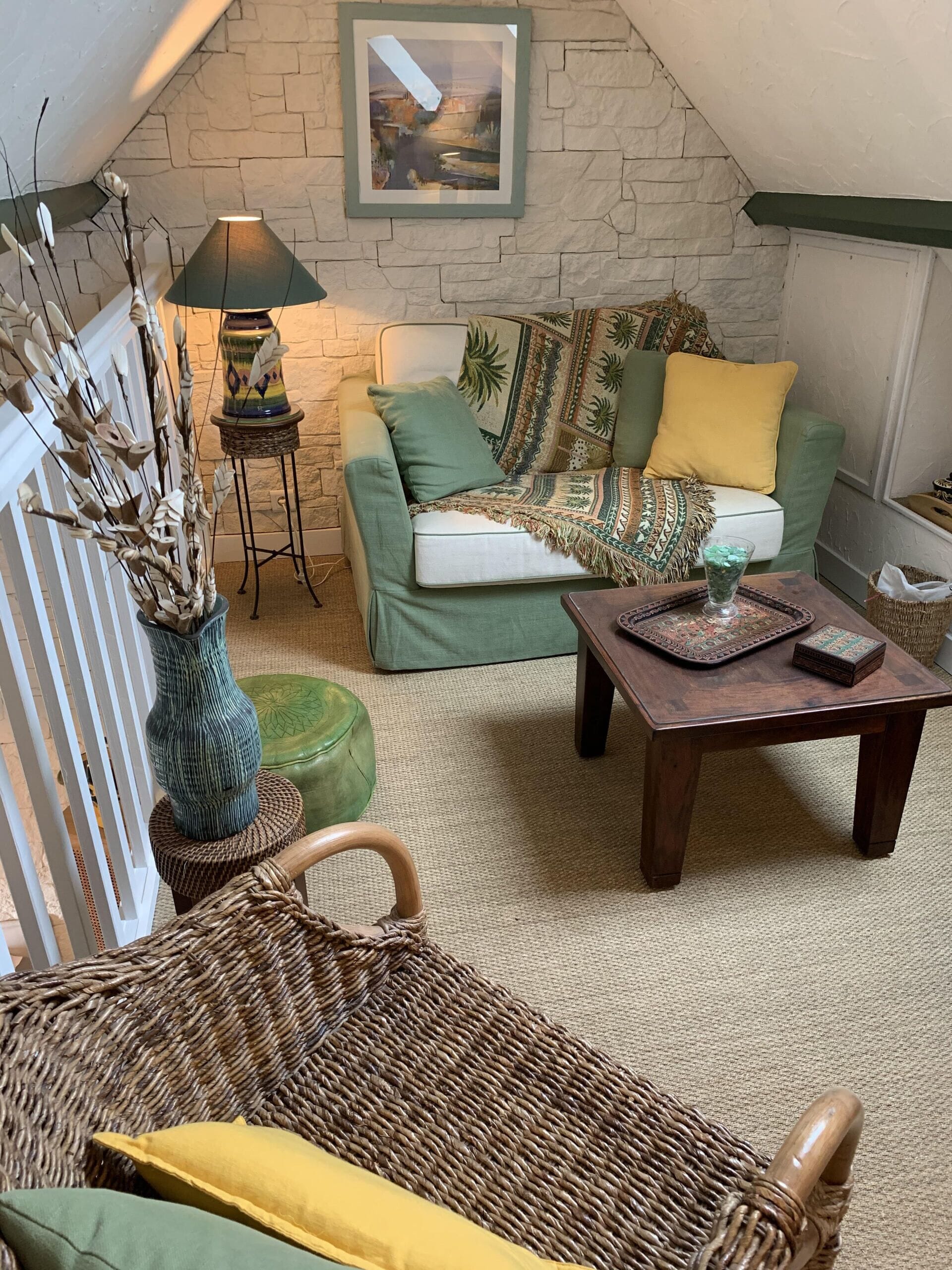 A cozy living room with a stone wall, a green sofa, a wicker chair, a wooden coffee table, and a lamp.