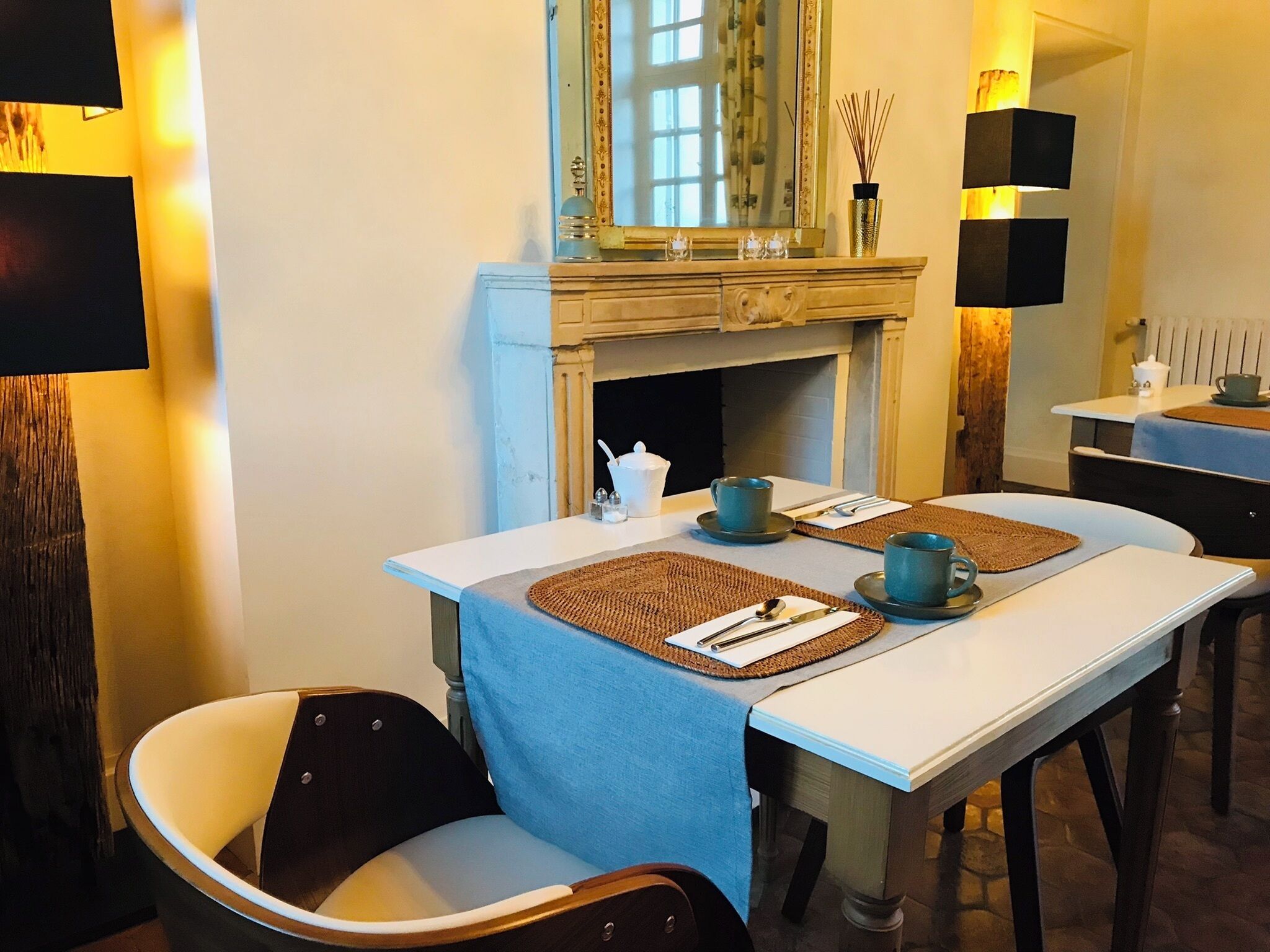 Un coin repas avec deux tables préparées pour deux personnes, chacune agrémentée de sets de table, de couverts et de tasses. Un miroir au-dessus de la cheminée reflète la pièce, et un vase contenant des roseaux est posé sur le manteau de la cheminée.