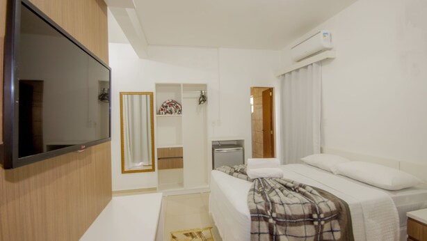 A hotel room with a flat-screen TV mounted on the wall, a bed with a plaid blanket, a small bedside table, and a white air conditioning unit on the wall. - Hotel Penha (Penha)