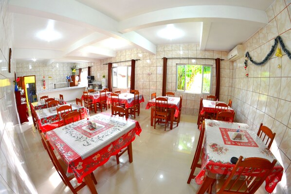 A dining area with multiple tables and chairs, a kitchen in the background, and a window with a view of greenery. - Hotel Penha (Penha)