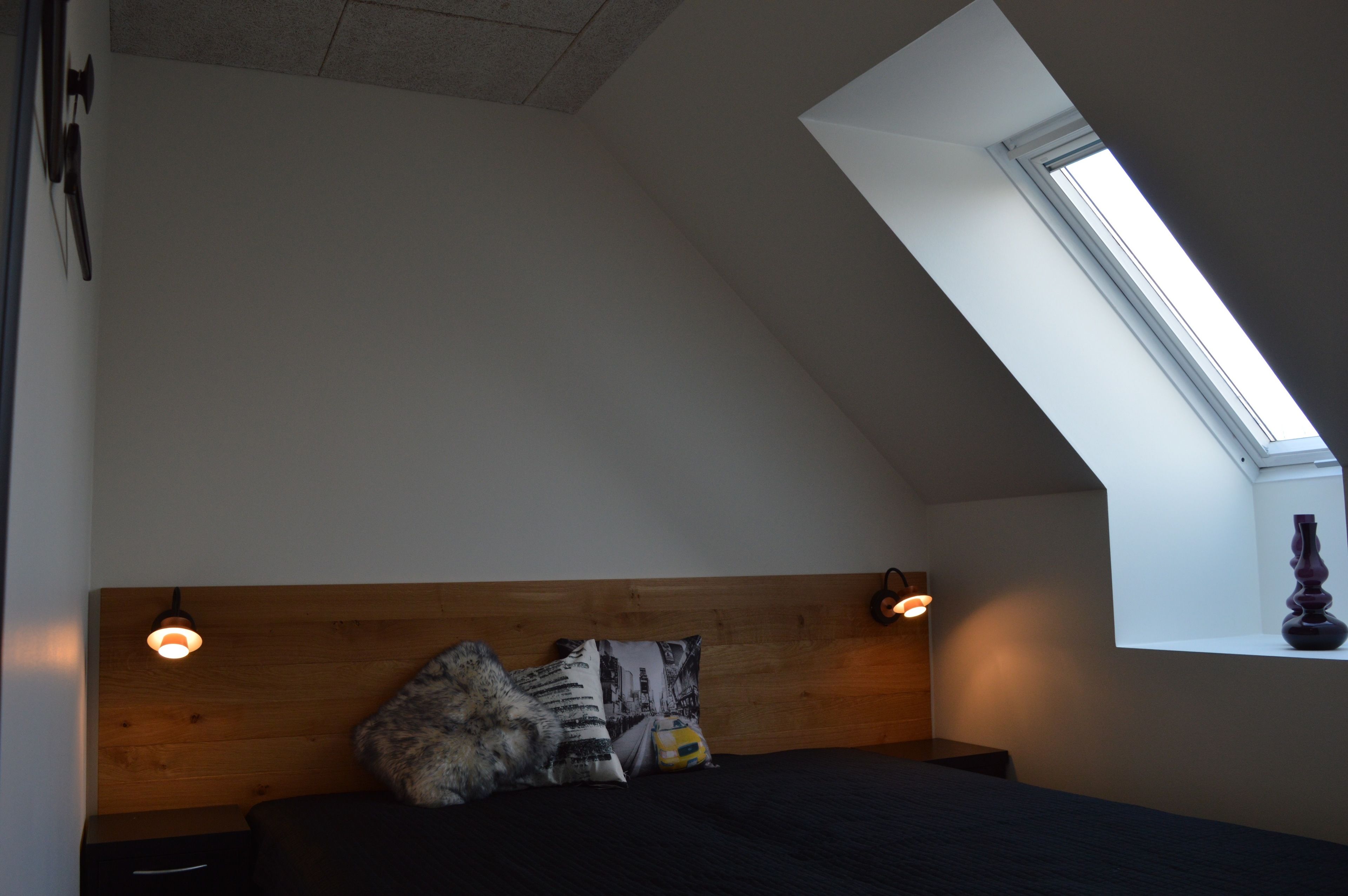 Un dormitorio con techo inclinado, tragaluces, cama con cabecera de madera y dos lámparas de noche.