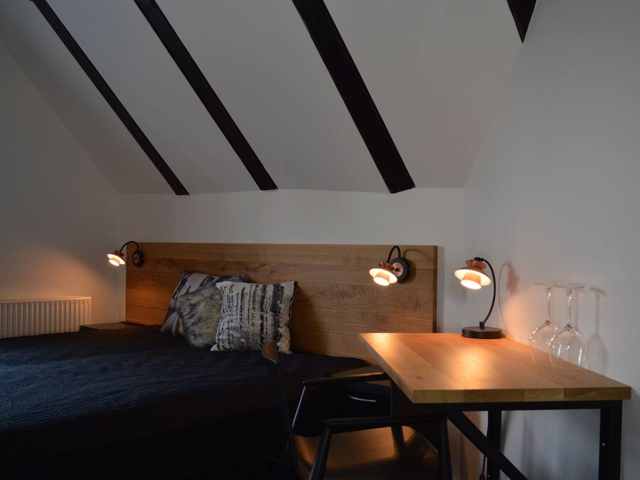A bedroom with a bed, a wooden headboard, two bedside lamps, a table with a chair, and a wine glass set on the table.