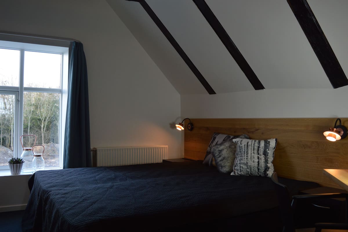 Un dormitorio con un ventanal grande, una cama con colcha oscura, dos lámparas de lectura fijadas a la pared y un cabecero de madera.