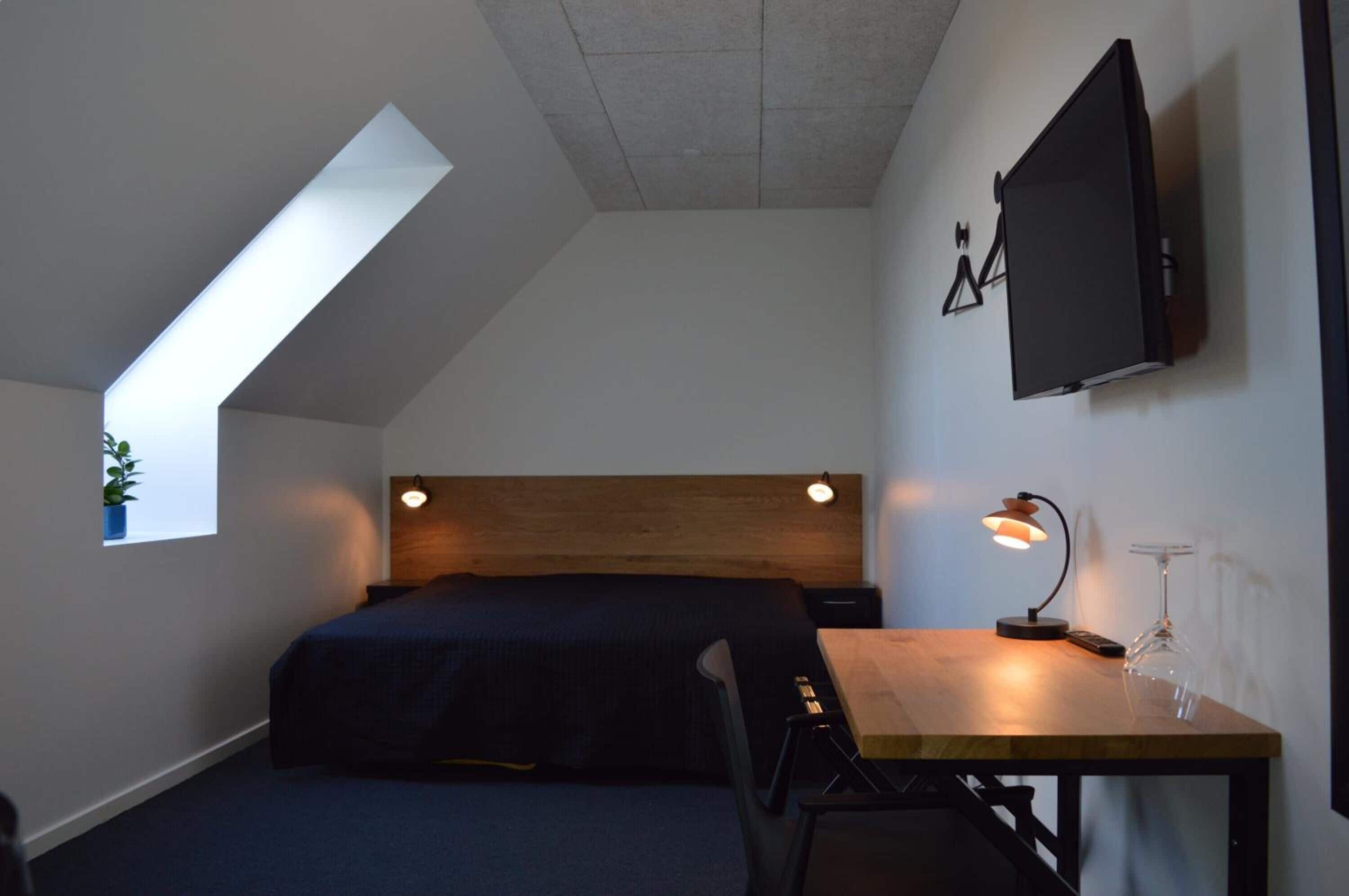Un dormitorio con techo inclinado, una cama, un escritorio con lámpara, un televisor montado en la pared y un ventanal con una planta.