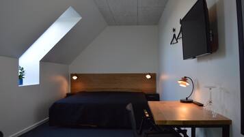 Un dormitorio con techo inclinado, una cama, un escritorio con lámpara, un televisor montado en la pared y un ventanal con una planta.