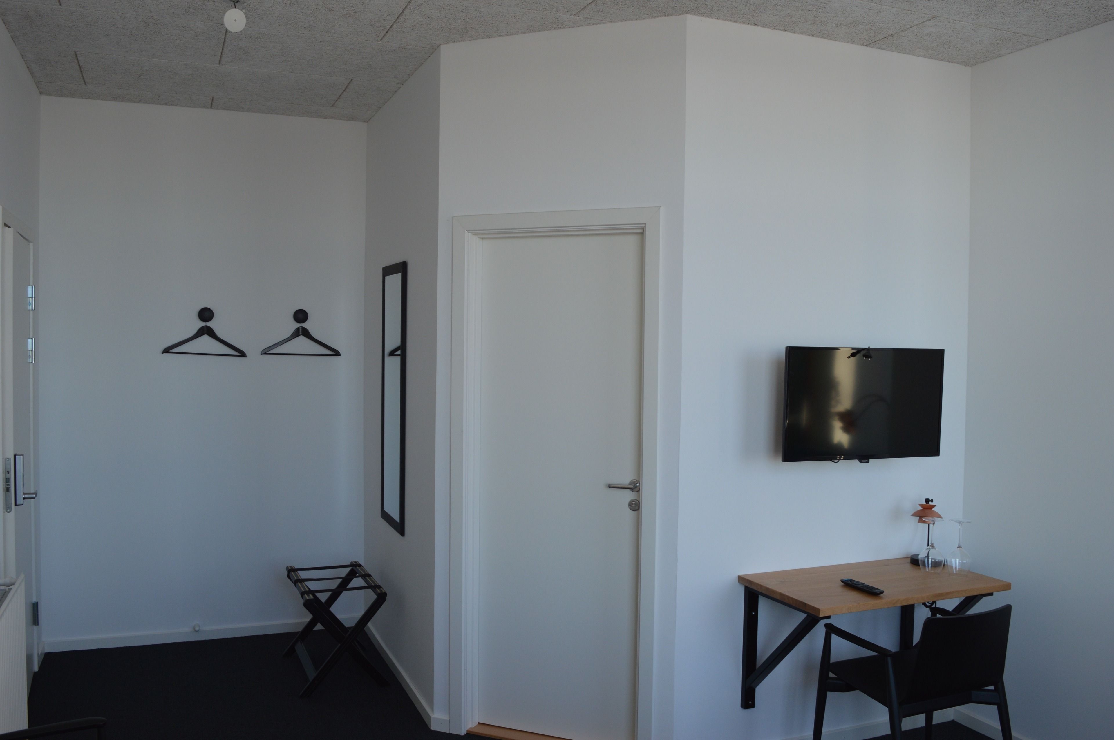 Una habitación con cama, escritorio con silla, televisor montado en la pared y una puerta.
