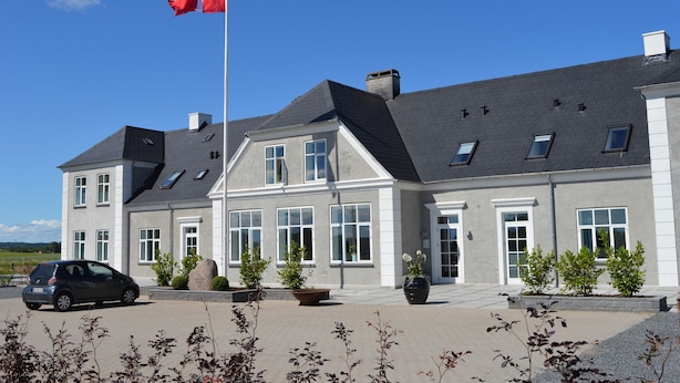 A two-story building with a grey roof and white walls, featuring multiple windows and a flagpole with a flag. - Femhøj (Jægerspris)