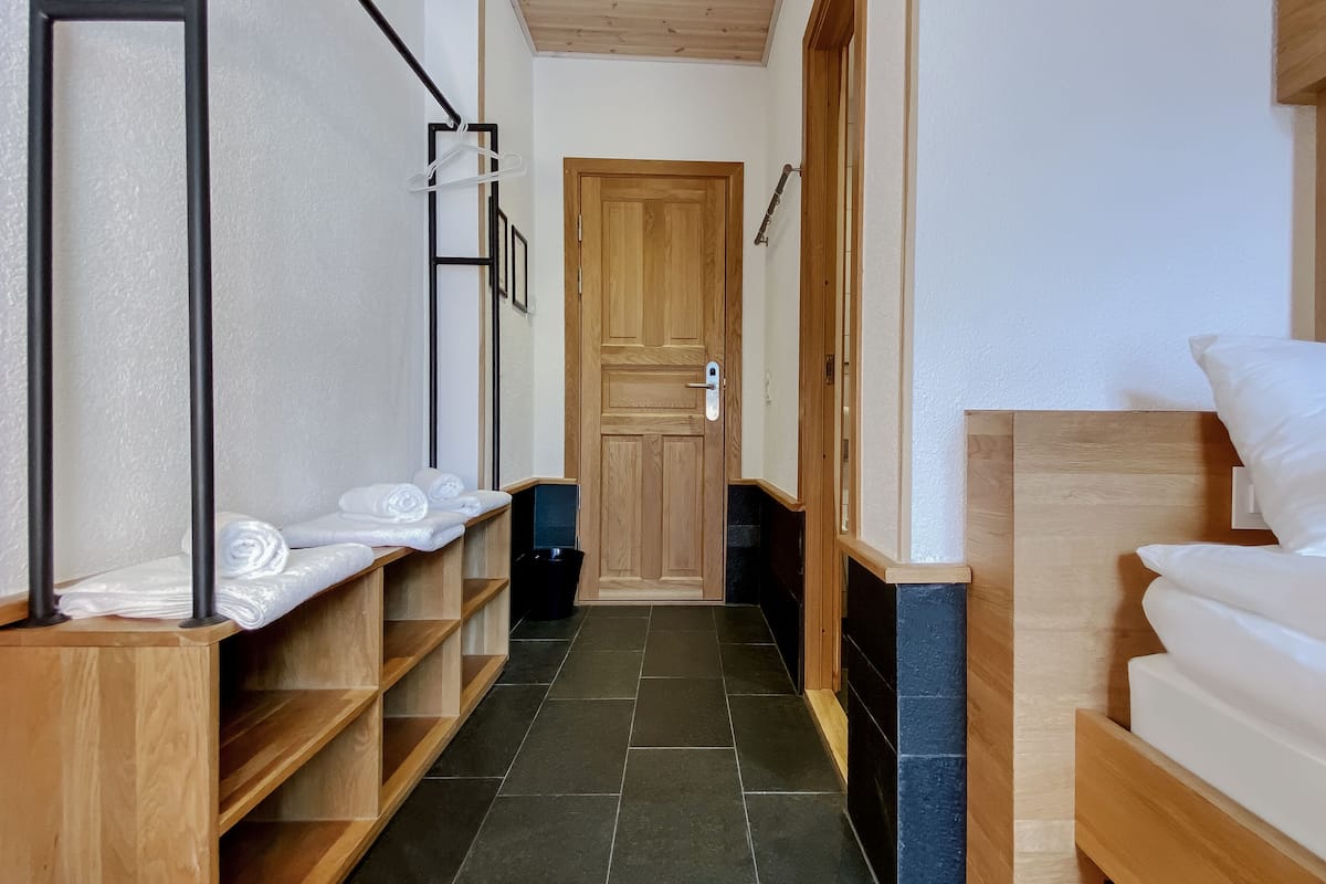 Una habitación de hotel con una cama de madera, un banco de madera con toallas, un cesto de basura negro y una puerta de madera.