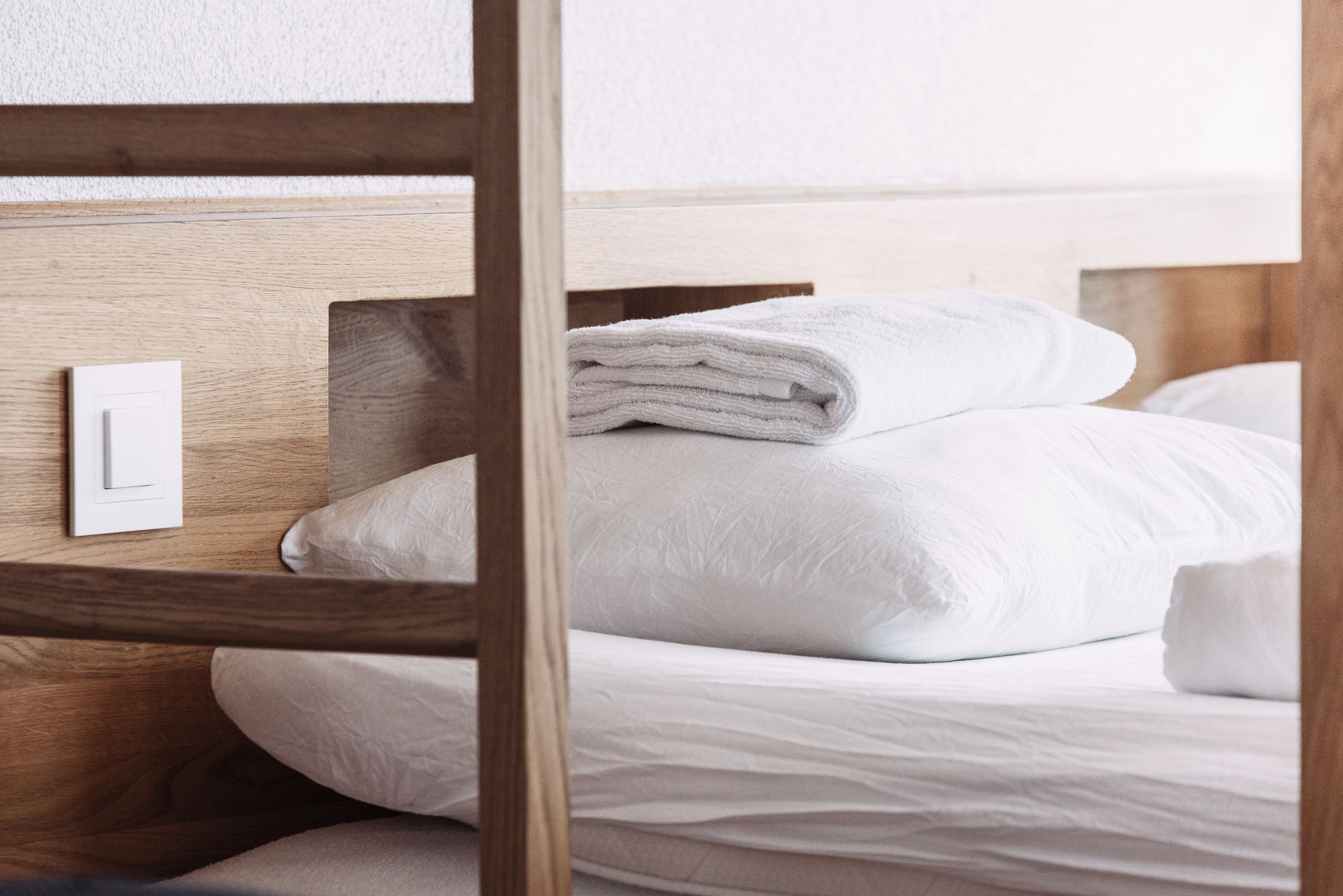 Una cama bien hecha con sábanas blancas y una toalla doblada encima, un cabecero de madera y un interruptor de luz en la pared.