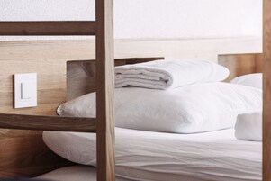A neatly made bed with white linens and a folded towel on top, a wooden headboard, and a light switch on the wall.