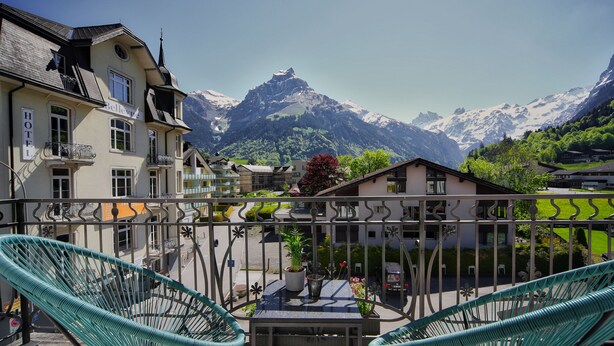 A hotel with a balcony featuring two green wicker chairs and a table, overlooking a mountain range. - GRAND your home with common kitchens with self check-in (Engelberg)