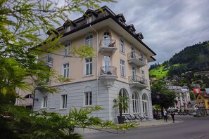 A multi-story building with balconies, a street in front, and a hilly background.