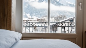 A bedroom with a large window offering a view of snow-covered mountains and a balcony with a metal railing. - GRAND your home with common kitchens with self check-in (Engelberg)