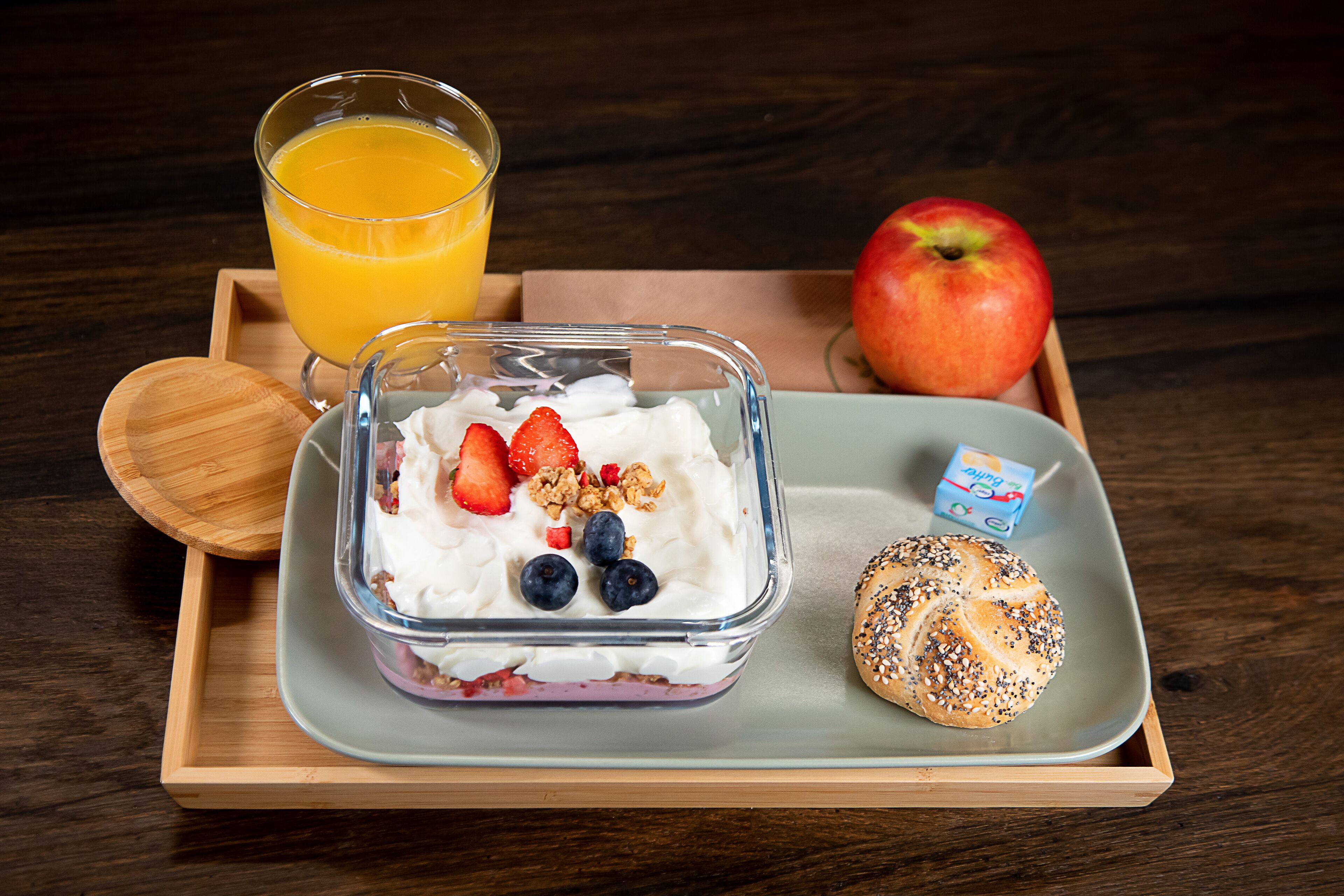 A breakfast tray with a glass of orange juice, a container of yogurt with strawberries and blueberries, a whole apple, a bagel with seeds, and a small carton of milk.