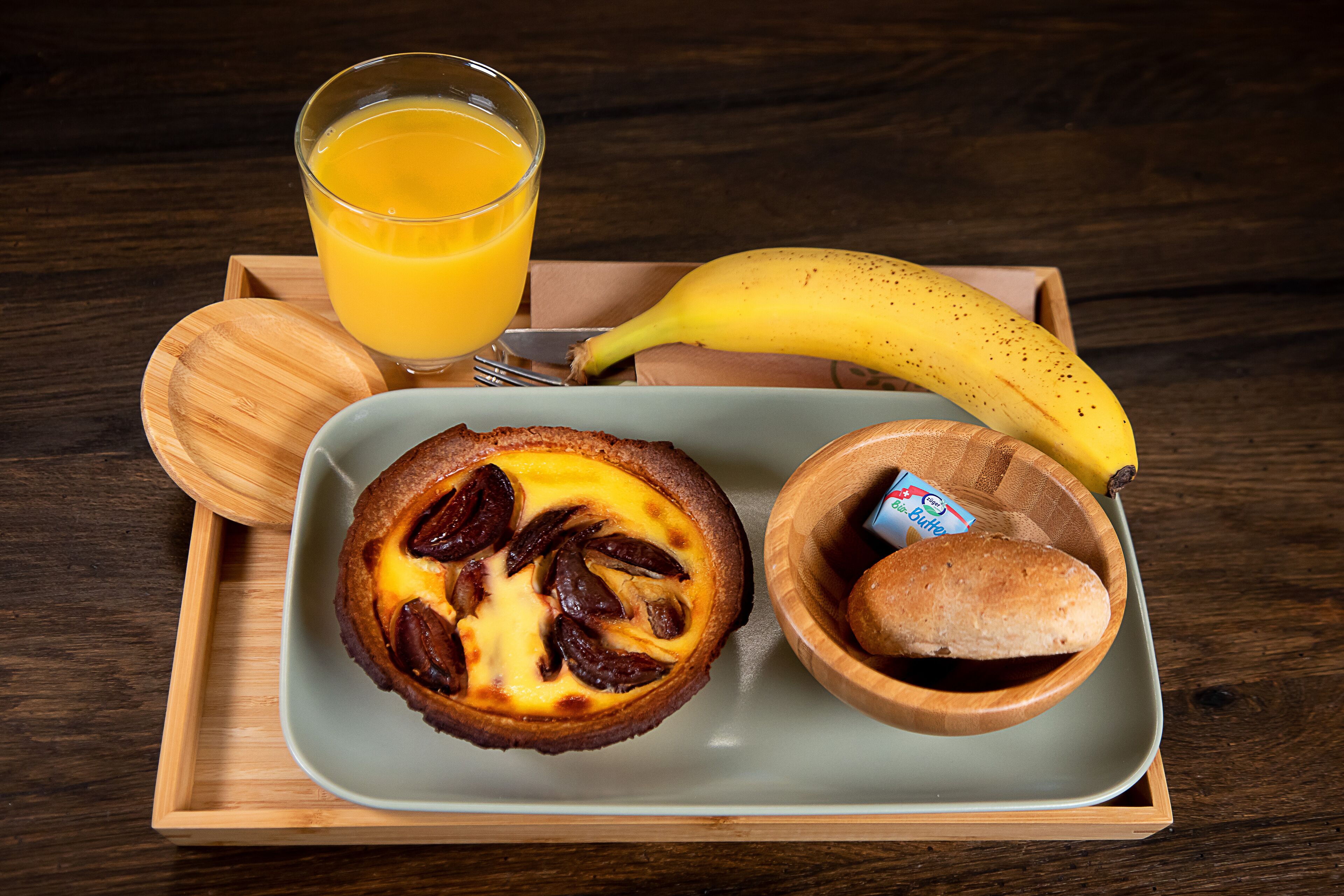 Bandeja de desayuno con una quiche, un vaso de jugo de naranja, un plátano, una rebanada de pan y un recipiente pequeño de mantequilla.