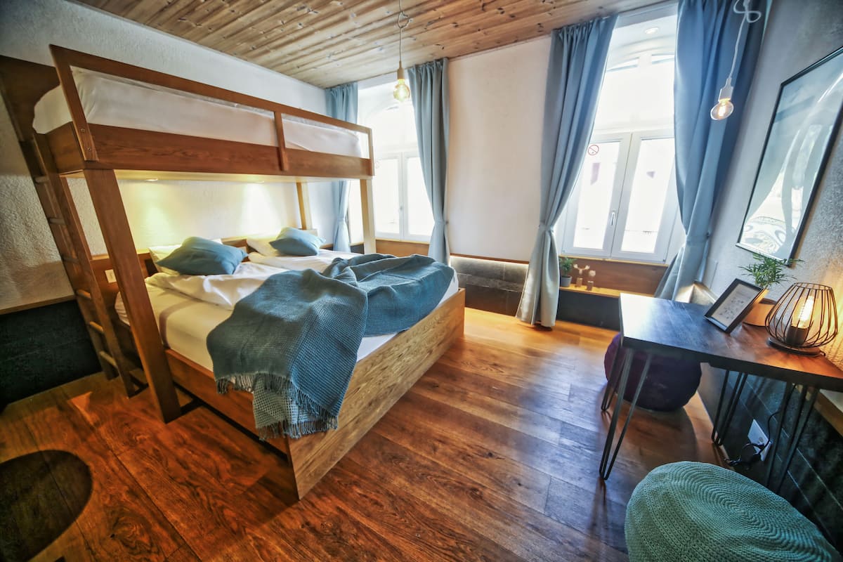 Una cama litera de madera con escritorio y silla, una ventana con cortinas y una lámpara.