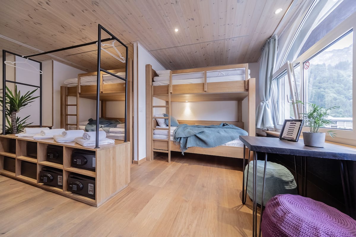 Habitación pequeña y bien iluminada con literas, un escritorio con una planta y un ventanal con vistas a la montaña.