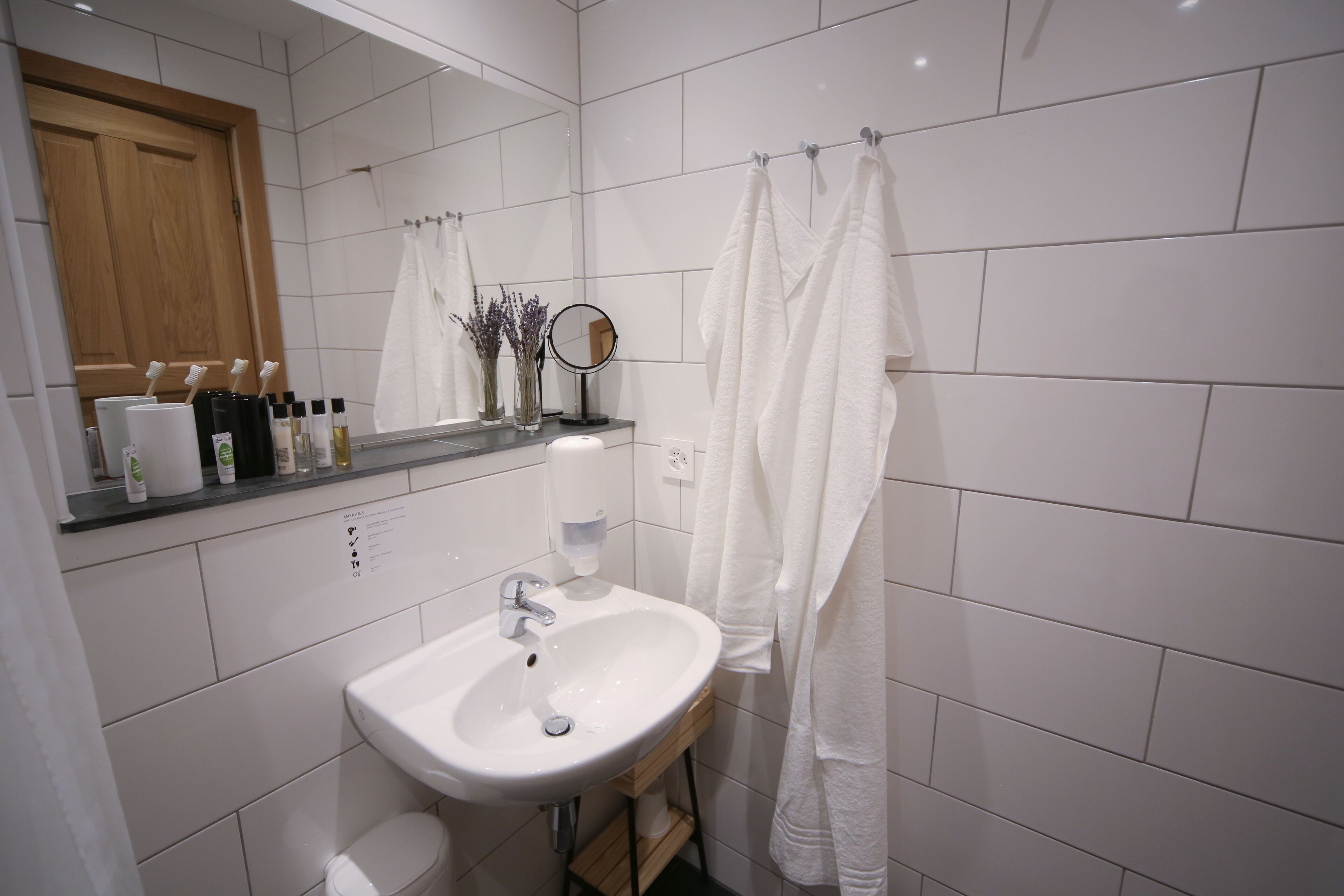 Un baño con lavamanos blanco, espejo y cortina de ducha. Hay artículos de aseo en la encimera y una puerta de madera al fondo.