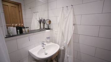 Un baño con lavamanos blanco, espejo y cortina de ducha. Hay artículos de aseo en la encimera y una puerta de madera al fondo.
