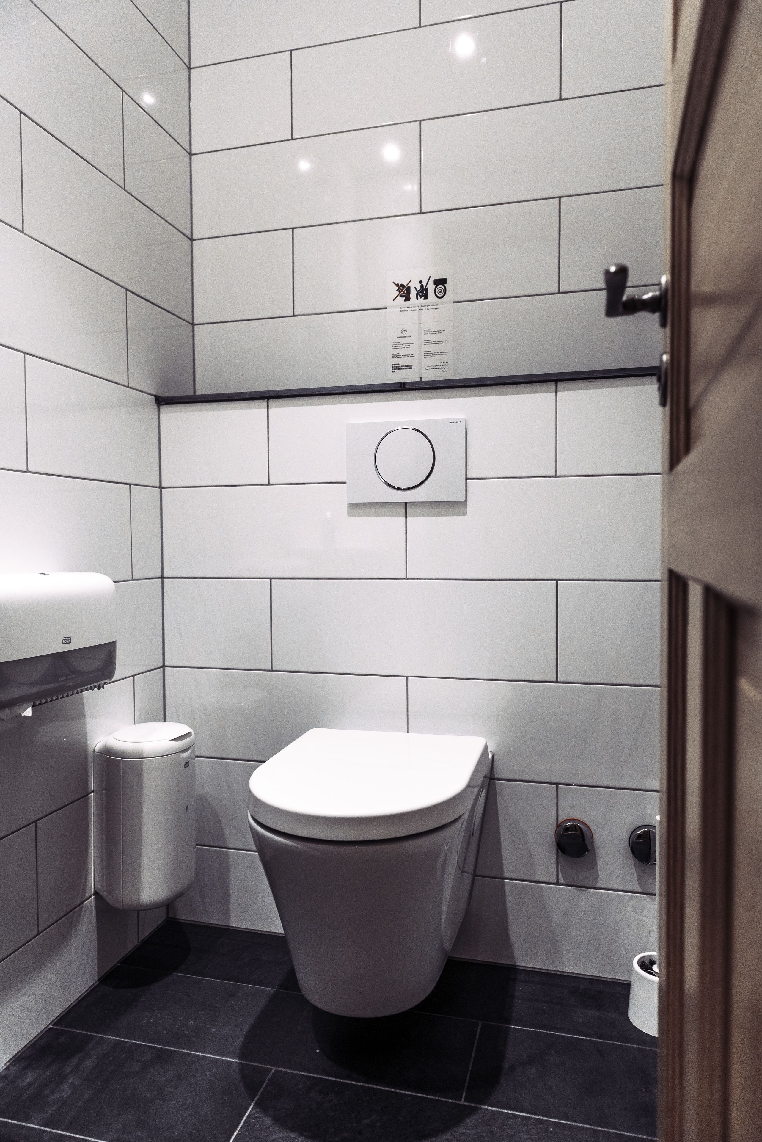 A modern bathroom with white tiles, a toilet, a trash bin, and a hand dryer.